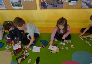 Dzieci siedzą na dywanie z rozłożonymi obrazkami, wybierają z pośród różnych herb Warszawy.